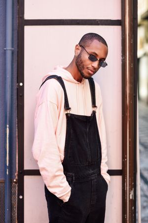 Male in denim overalls and earrings with hands in pockets against a pink wall