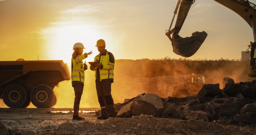 Cinematic Golden Hour Shot Of Construction Site: Male Civil Engineer And Hispanic Female Urban Planner Talking, Using Tablet. Trucks, Excavators, Loaders Working To Build New Apartment Block