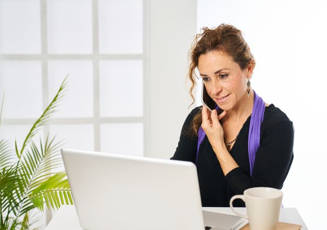 a woman smiling while answering a phone call and typing on a laptop