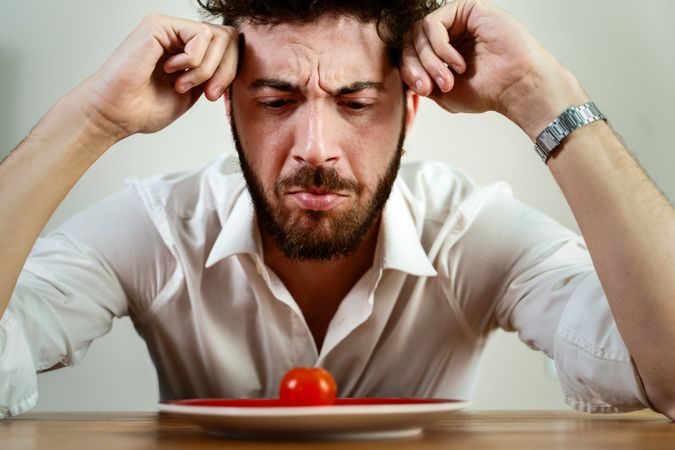 Frustrated man on a strict diet looking at a single cherry tomato on a plate.
