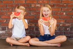 Cute Young Boy and Girl Eating Watermelon Against Brick Wall
