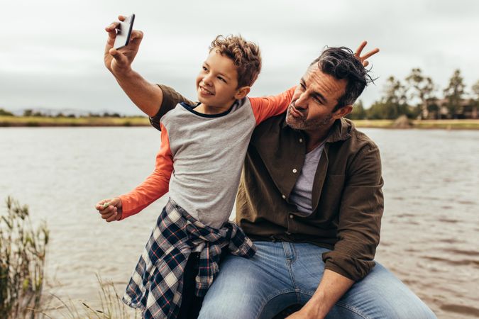 Man sitting on a tree trunk beside a lake with his son and taking a selfie