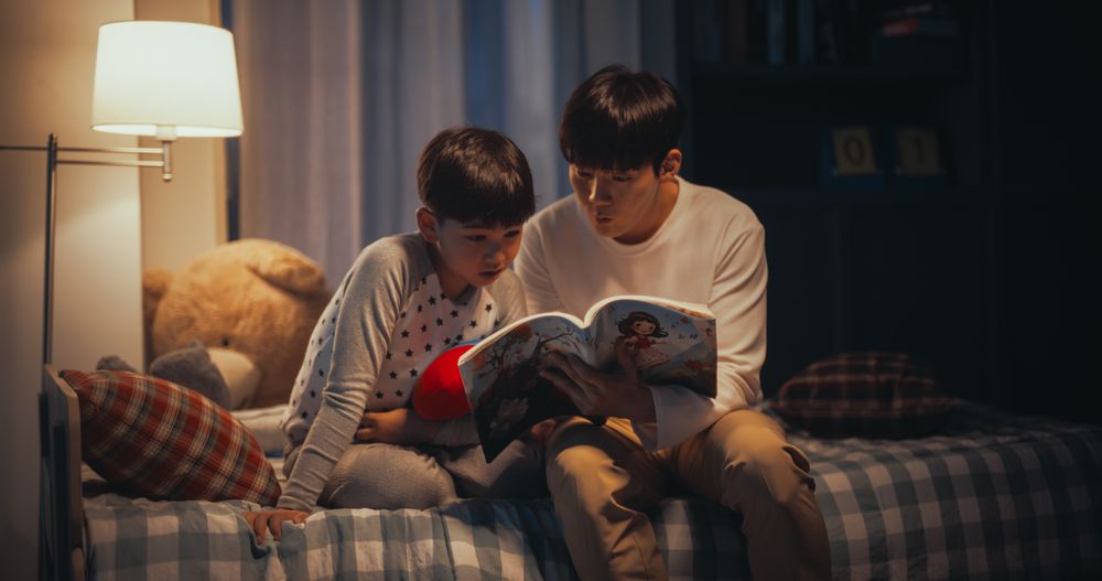 Korean Father Reading a Fairytale to His Lovely Cute Son in Bed Before Going to Sleep.