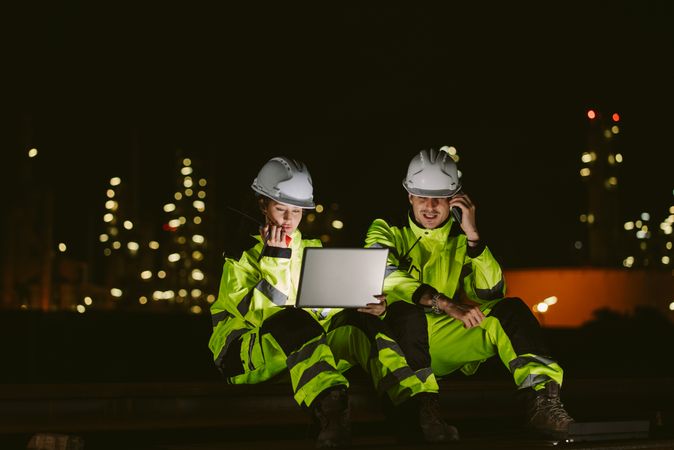 Team of two industrial workers using laptop, walkie-talkie, and phone at night