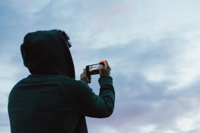 Rearshot of man wearing hood on cliff taking photo on phone at sunset