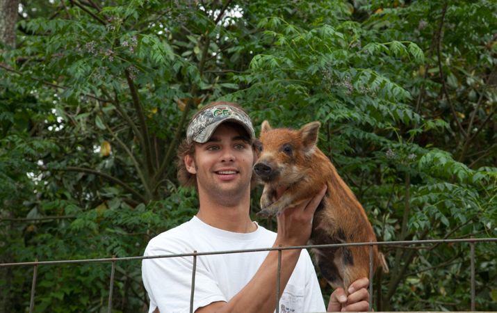 Young white man in visor holds red furry piglet on Meadowbank Farm in Alabama