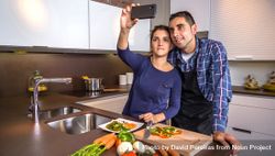 Content couple in a home kitchen taking a selfie with a smartphone while preparing meal bGjml4