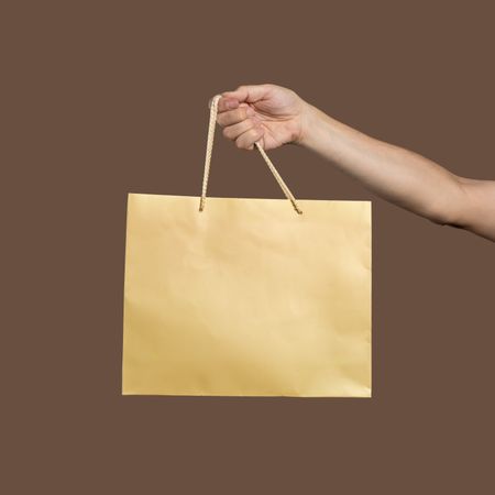 Hand holding a blank gold shopping bag mockup on a brown background.