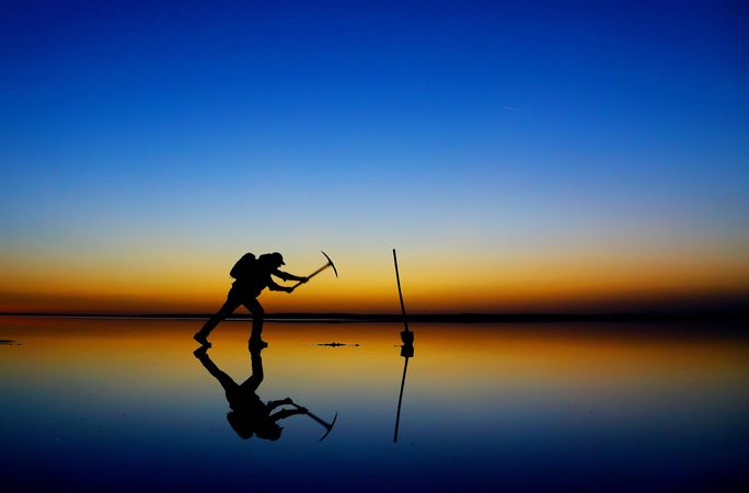 Silhouette of a man digging a hole in a frozen lake during sunset