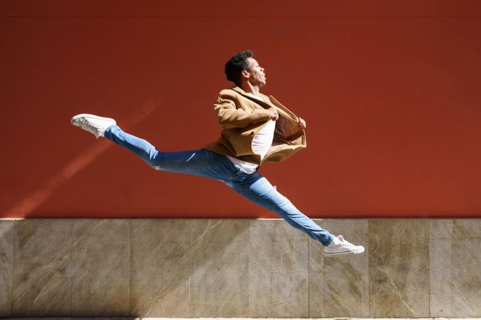 Graceful Black male dancer in camel coat jumping against am outdoor wall