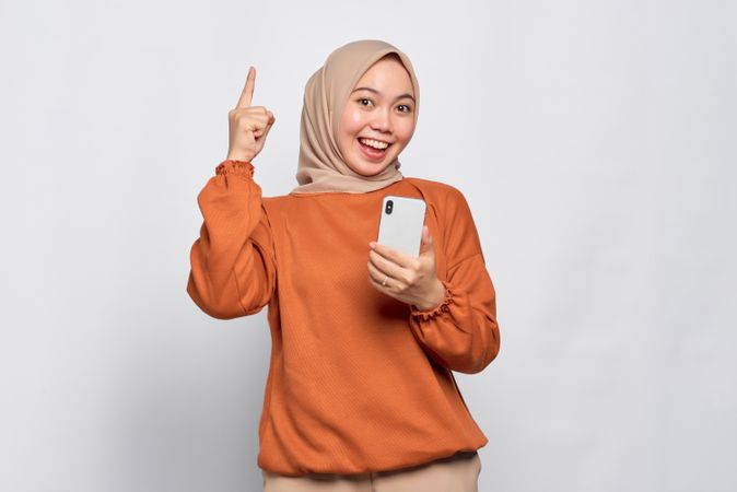 Muslim woman smiling holding smart phone and pointing fingers up with a bright idea