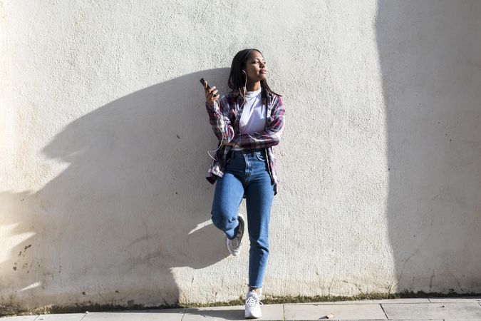 Female standing in the sun while leaning on wall listening to something on smartphone