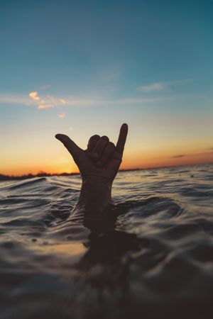Hand with shaka gesture between water surface and sky at sunset
