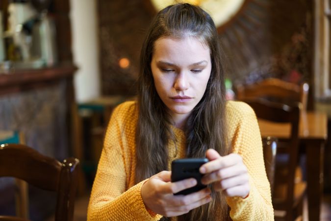 A young woman is deeply engrossed in her smartphone while sitting inside a cozy cafe