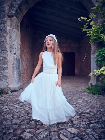Young girl in long white dress and wreath standing near old buil