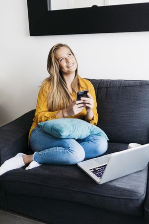 Dreamy long haired female sitting on sofa with laptop computer working remotely from home checking phone