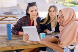 Three friends on park bench with insulated mugs looking at something on digital tablet