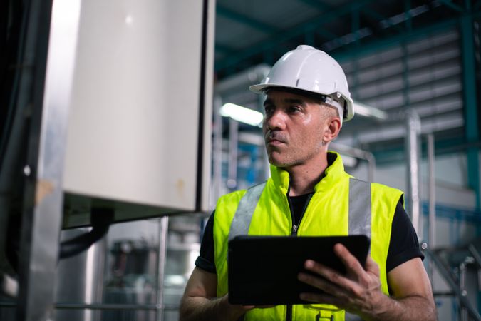 A male industrial engineer wearing a safety helmet and reflective vest is inspecting equipment in a factory. He is using a digital tablet to review data. Background show industrial pipes and machinery