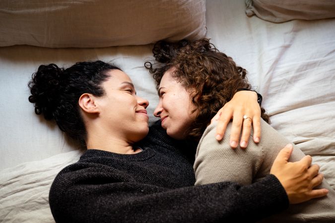 Two women lying down on bed and holding each other looking at each other with tenderness