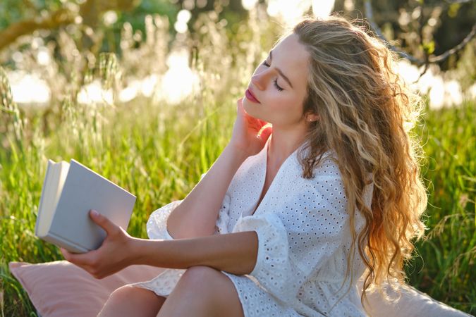 Dreamy woman with book touching neck gently in lush park