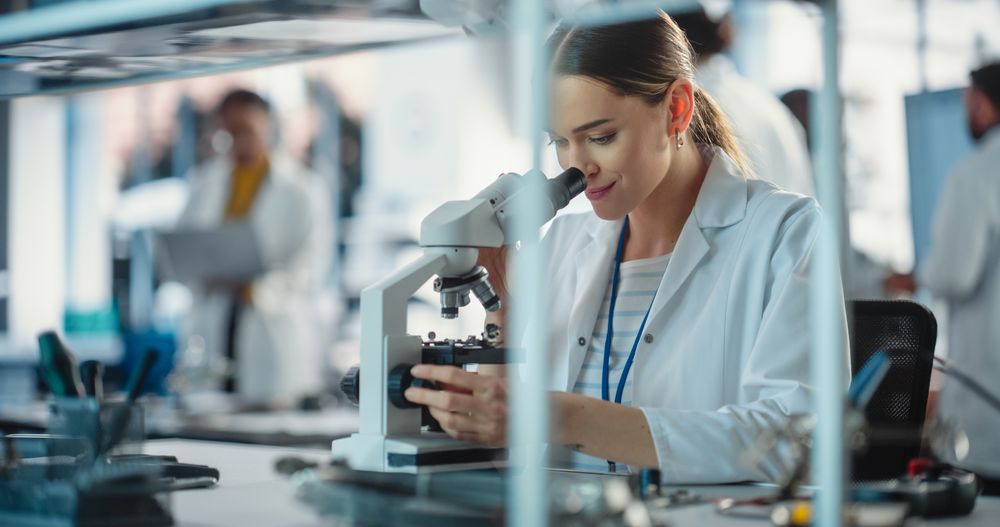 Project Managers Discuss Manufacturing Using Laptop Computer While Female Specialist Checking on Electronic Circuit Using Microscope
