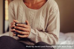 Close up of woman wearing sweater holding a cup of coffee while sitting on bed 5nG2m4