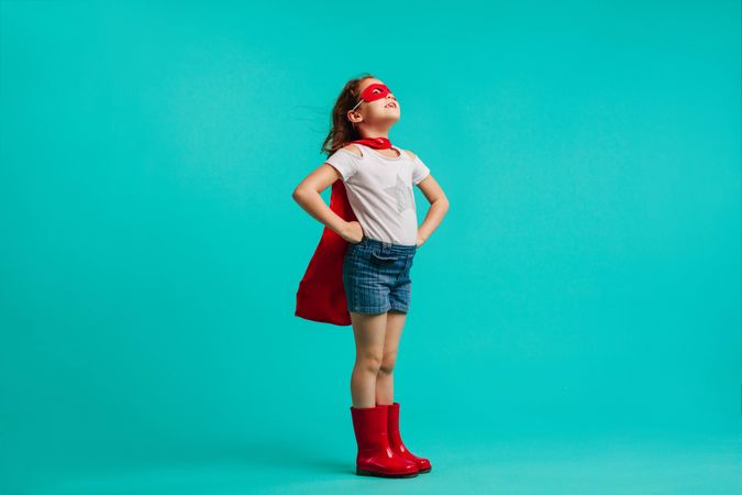 Girl wearing red gumboots, cape and eye mask standing with her hands on hips in studio