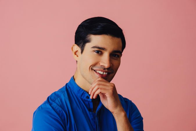 Hispanic man smiling at camera in pink studio with hand on chin