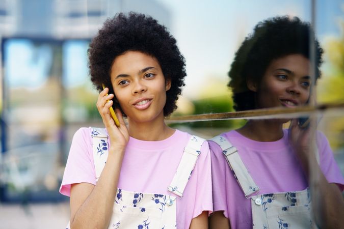 Female in floral overalls speaking on cell phone and leaning on window outside