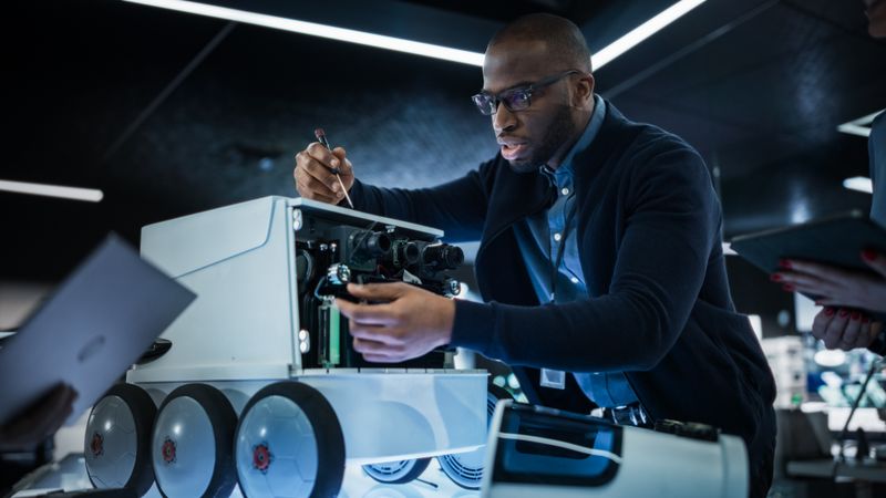 Engineer Using a Screwdriver While Developing a Remotely Controlled Delivery Robot. Black Specialist Working in High Tech Research Laboratory with Modern Equipment