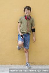 Young teen boy leaning on a yellow wall with headphones on neck bDjQA8