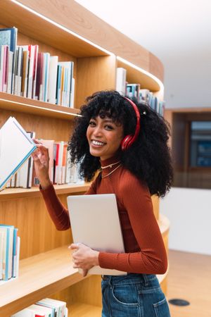 Cheerful female with laptop choosing book in library