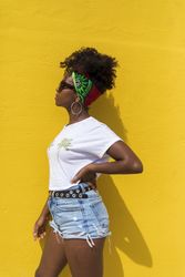 Side view of Black woman with sunglasses and headscarf leaning on yellow wall while looking away
