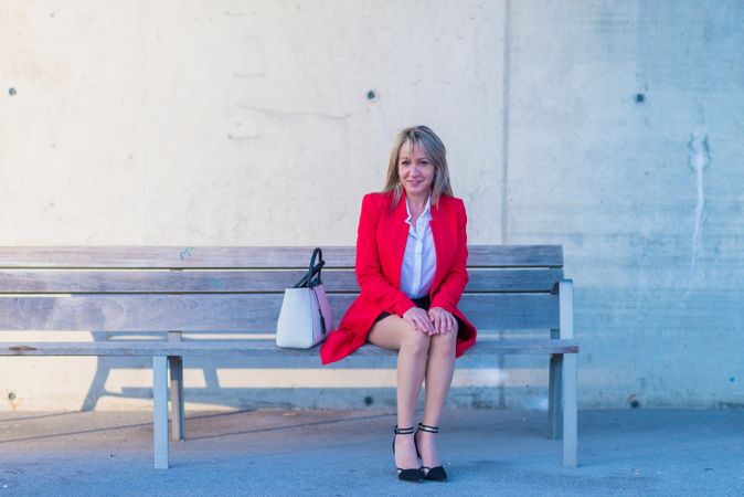 Blonde executive wearing red jacket sitting on a city bench while looking to camera and smiling
