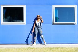 Female in denim overalls fixing sunglasses in front of blue building