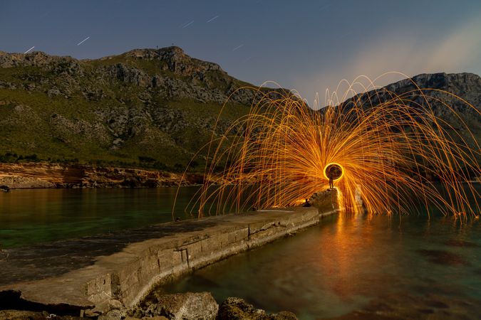 Person with burning circle on stones between water near hills at night
