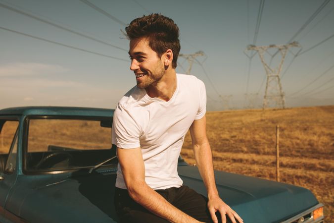 Happy man sitting on hood of truck parked during road trip to country side