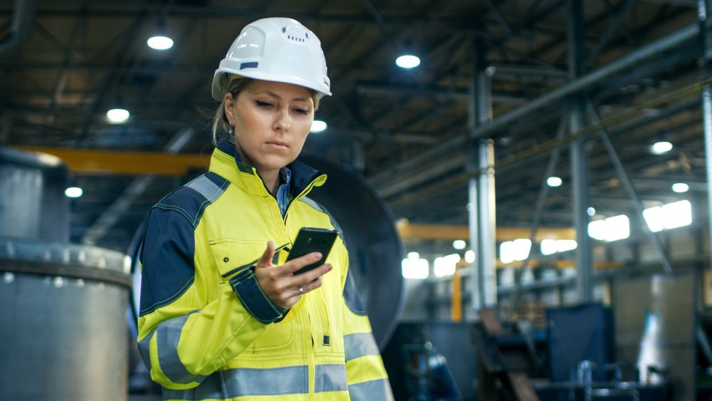 Female Industrial Worker in the Hard Hat Uses Mobile Phone While Walking Through Heavy Industry Manufacturing Factory.