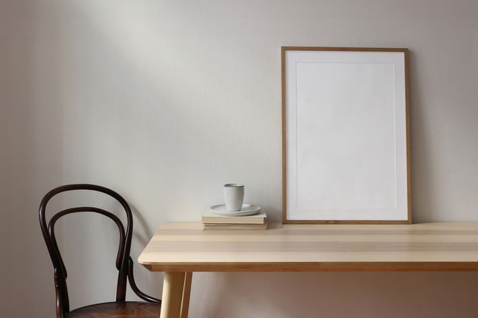 Empty wooden picture frame mockup. Cup of coffee, books on wooden table. Beige wall background in sunlight. Old chair. Working space, home office. Art, poster display. Elegant Scandi interior.