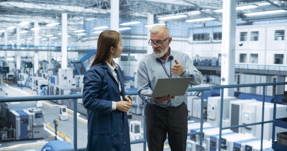 Two Diverse Engineers Standing on a Platform, Using Laptop Computer at an Electronics Factory. Machines are Producing Electronic Chips, Specialists Monitoring the Progress on Online Software