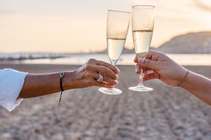 Crop women clinking wineglasses on beach during sunset