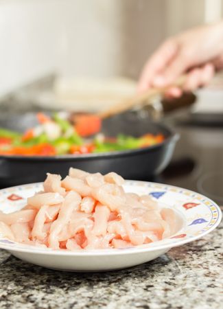 Closeup of raw chiken dish and female cooking in a pan in the background