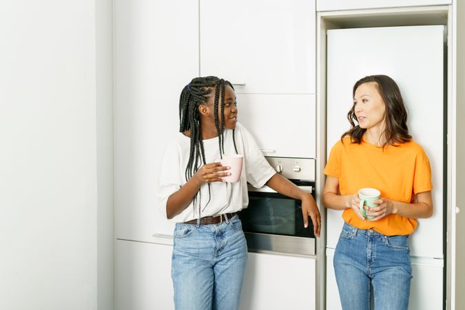 Two women chatting together while leaning on the fridge with coffee