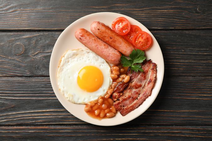Wooden table with breakfast plate of eggs, tomatoes, sausage and bacon, top view
