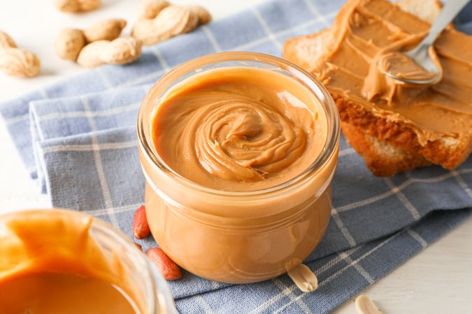 Glass jar with peanut butter, peanut, kitchen towel, spoon and peanut butter sandwich on wooden background, space for text and closeup