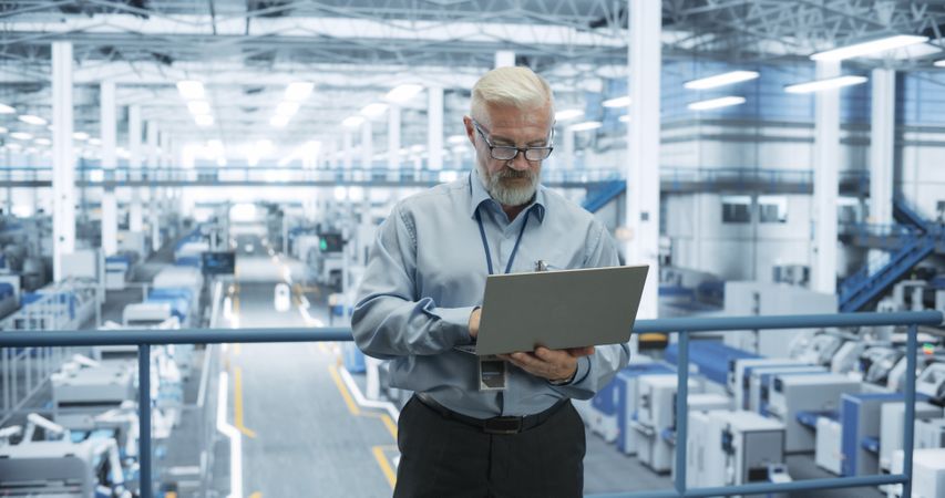 Engineer Standing on a Platform, Using Laptop Computer at an Electronics Factory. Machines are Undergoing Maintenance, Specialist Monitoring the Updates Through Online Software