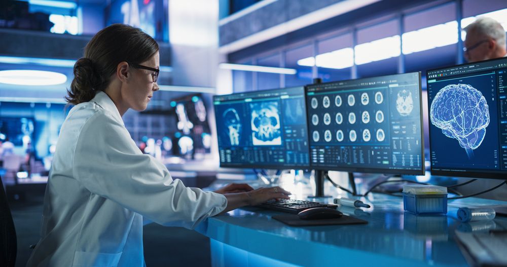 White Female Neuroscientist Using Desktop Computer To Analyze CT Scans Of Human Brain In Busy Research Center Laboratory.