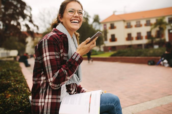 Smiling woman sitting at university campus with book talking on mobile phone