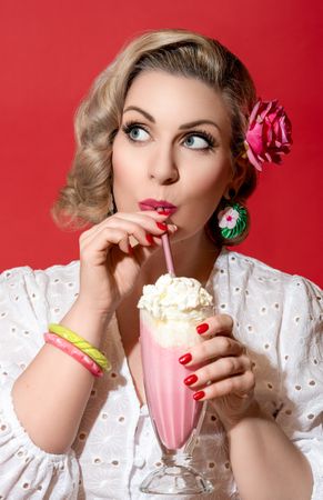 Blonde woman with a retro hairstyle and clothes drinking a strawberry milkshake in red studio
