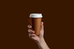 Hand holding a takeaway paper coffee cup on a solid brown background.